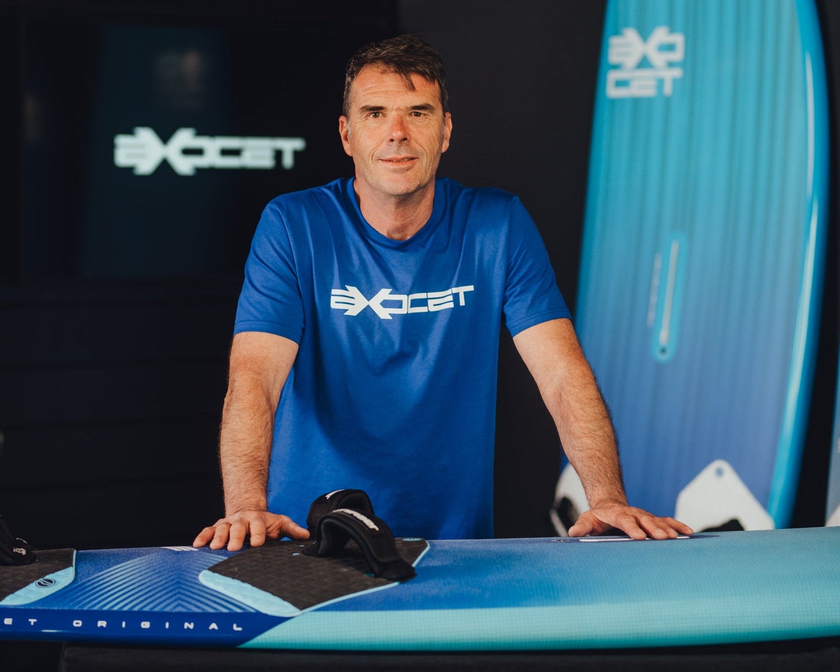 Man in a blue shirt standing next to a blue windsurf board with Exocet branding.